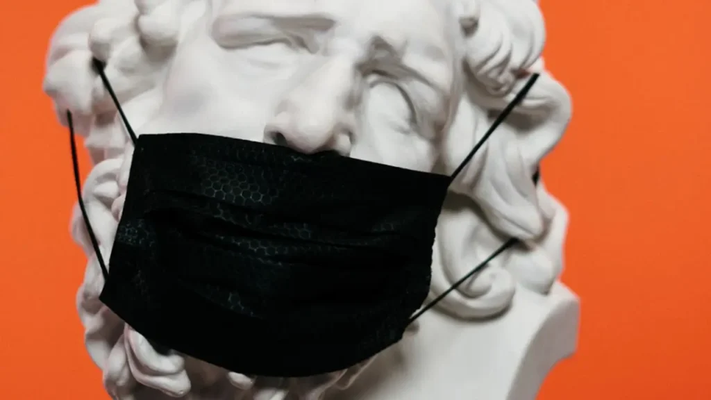 Une statue en marbre blanc représentant une personne aux cheveux bouclés porte un masque noir. Le fond orange crée un contraste saisissant avec la statue blanche et le masque sombre.